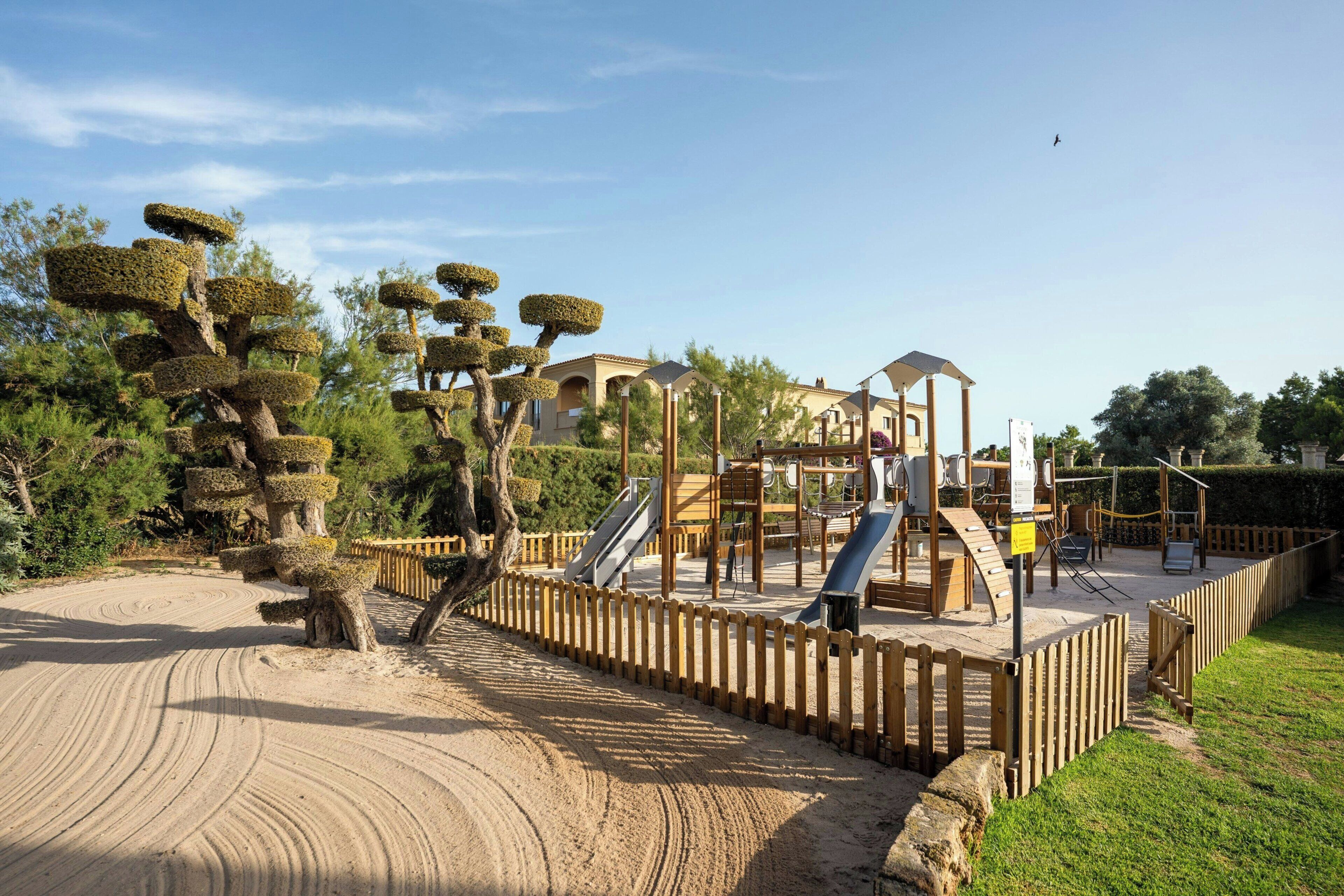 Un parque infantil con tobogán y estructuras de escalada, rodeado por una cerca de madera y una zona de arena.