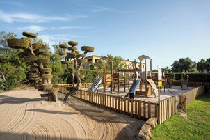 Un parque infantil con tobogán y estructuras de escalada, rodeado por una cerca de madera y una zona de arena.