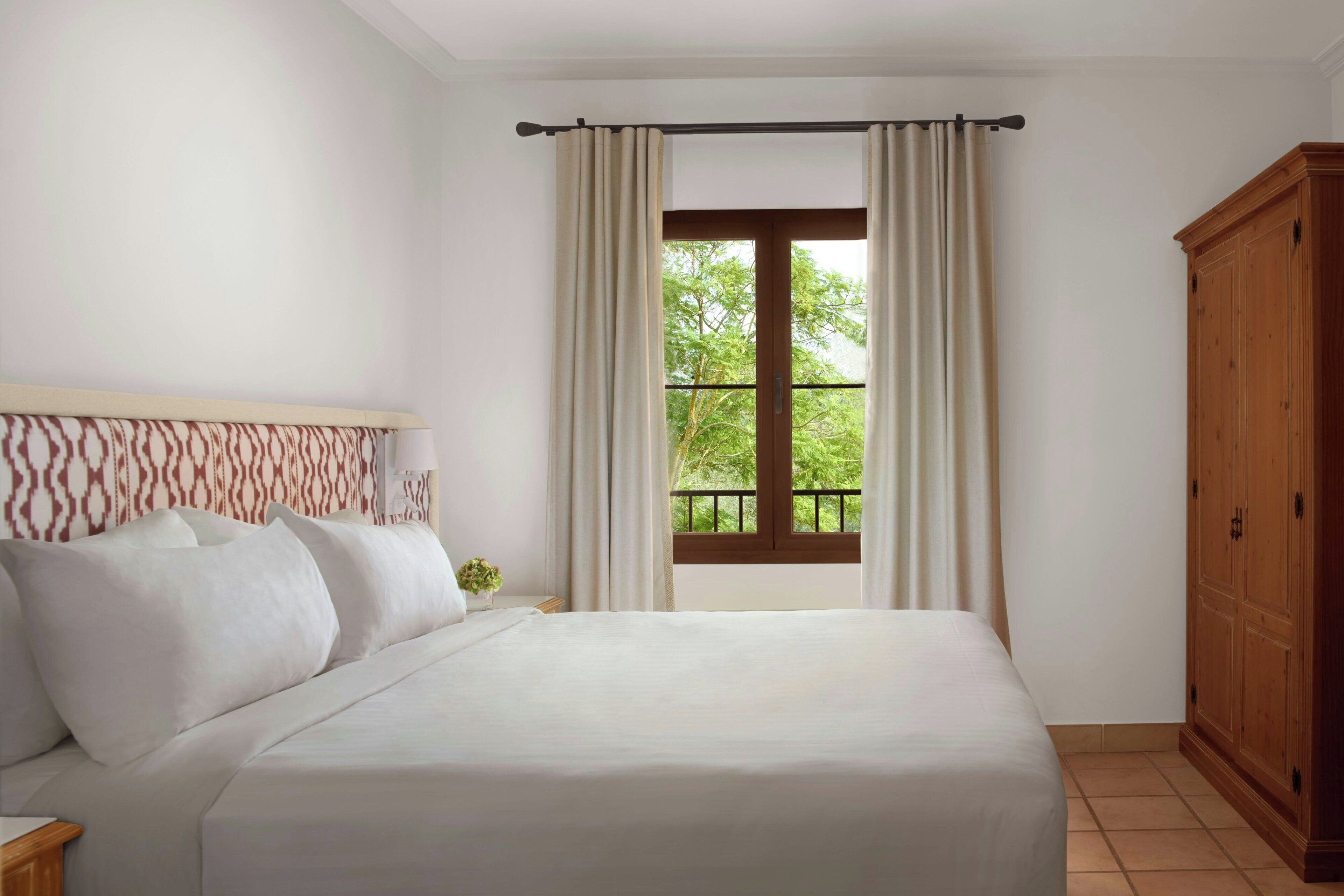 Une chambre à coucher avec un lit, une armoire en bois, une fenêtre avec des rideaux et une vue sur la verdure.