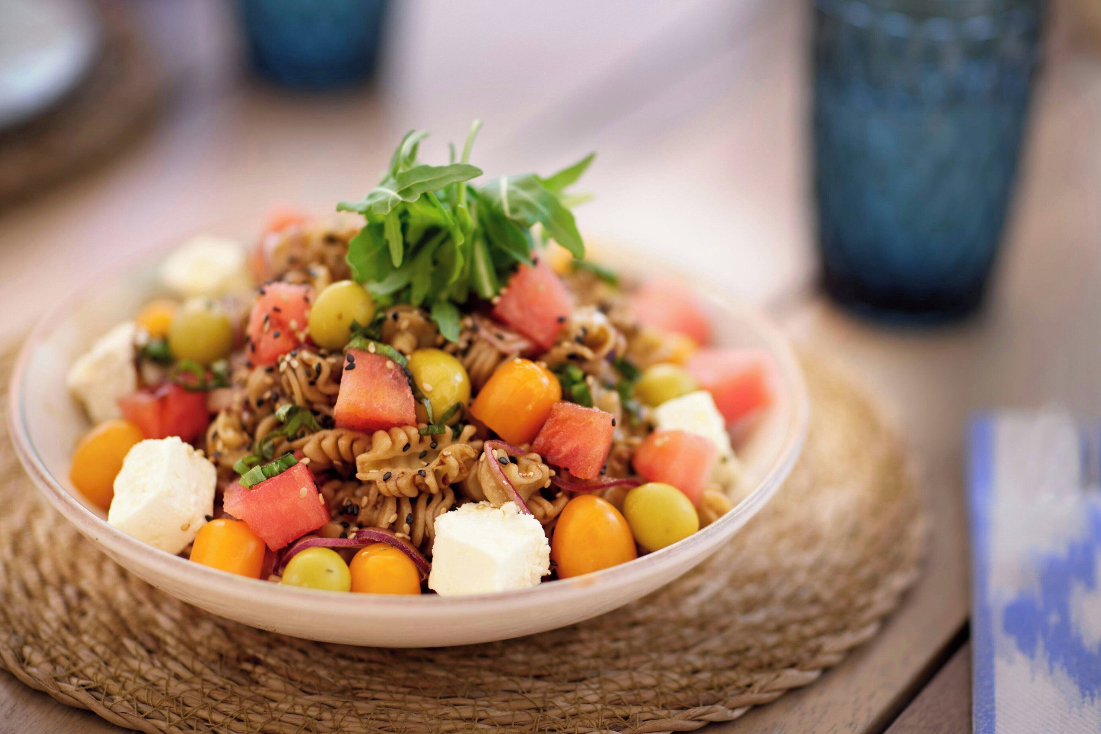 Un tazón de ensalada de pasta con diferentes verduras y queso, sobre una estera trenzada.
