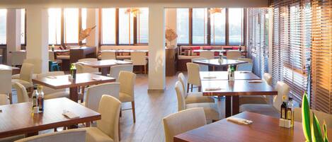 A hotel dining area with multiple tables and chairs, large windows with blinds, and a modern interior design.