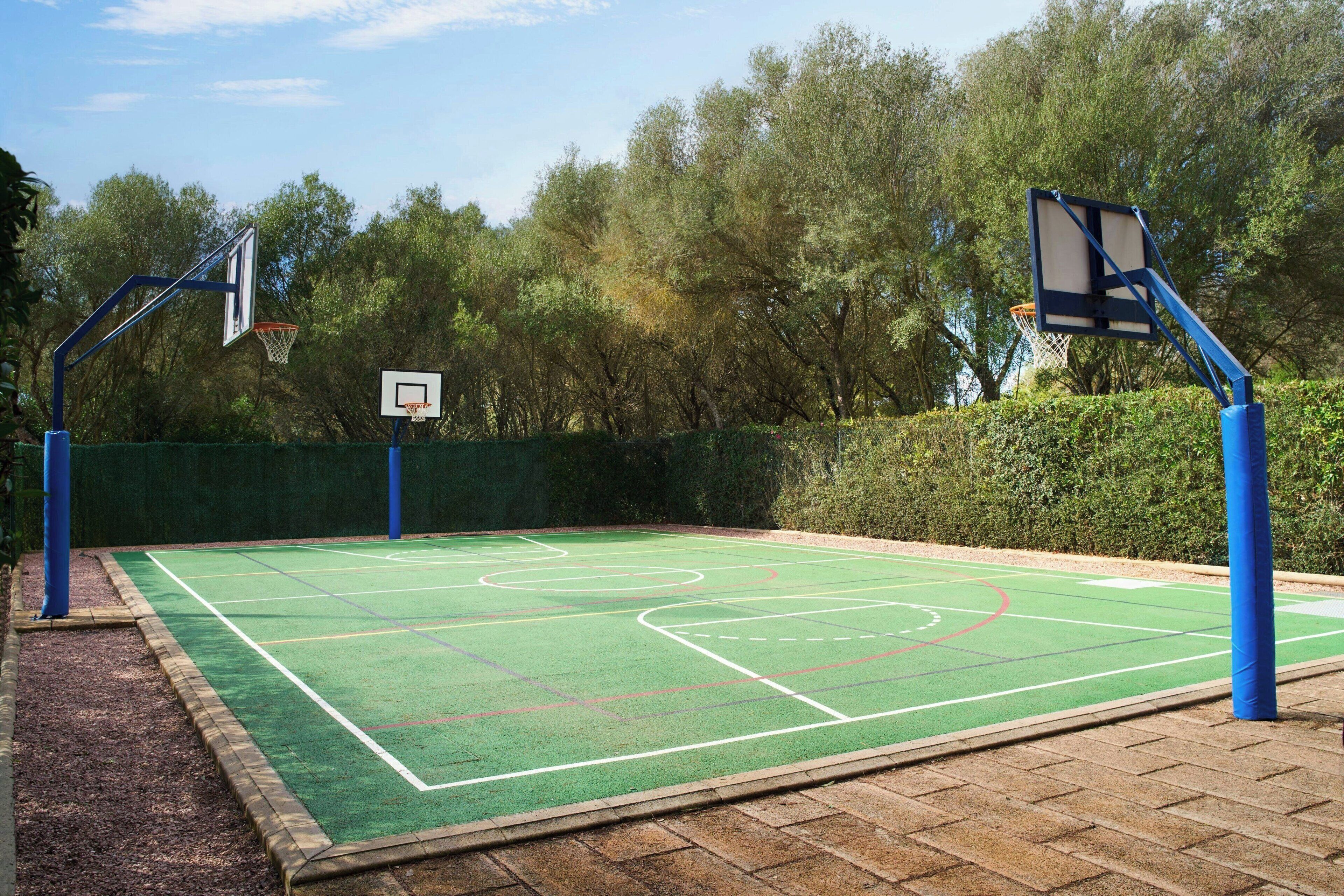 Quadra de basquete ao ar livre com duas cestas, cercada por árvores e um muro de sebe.