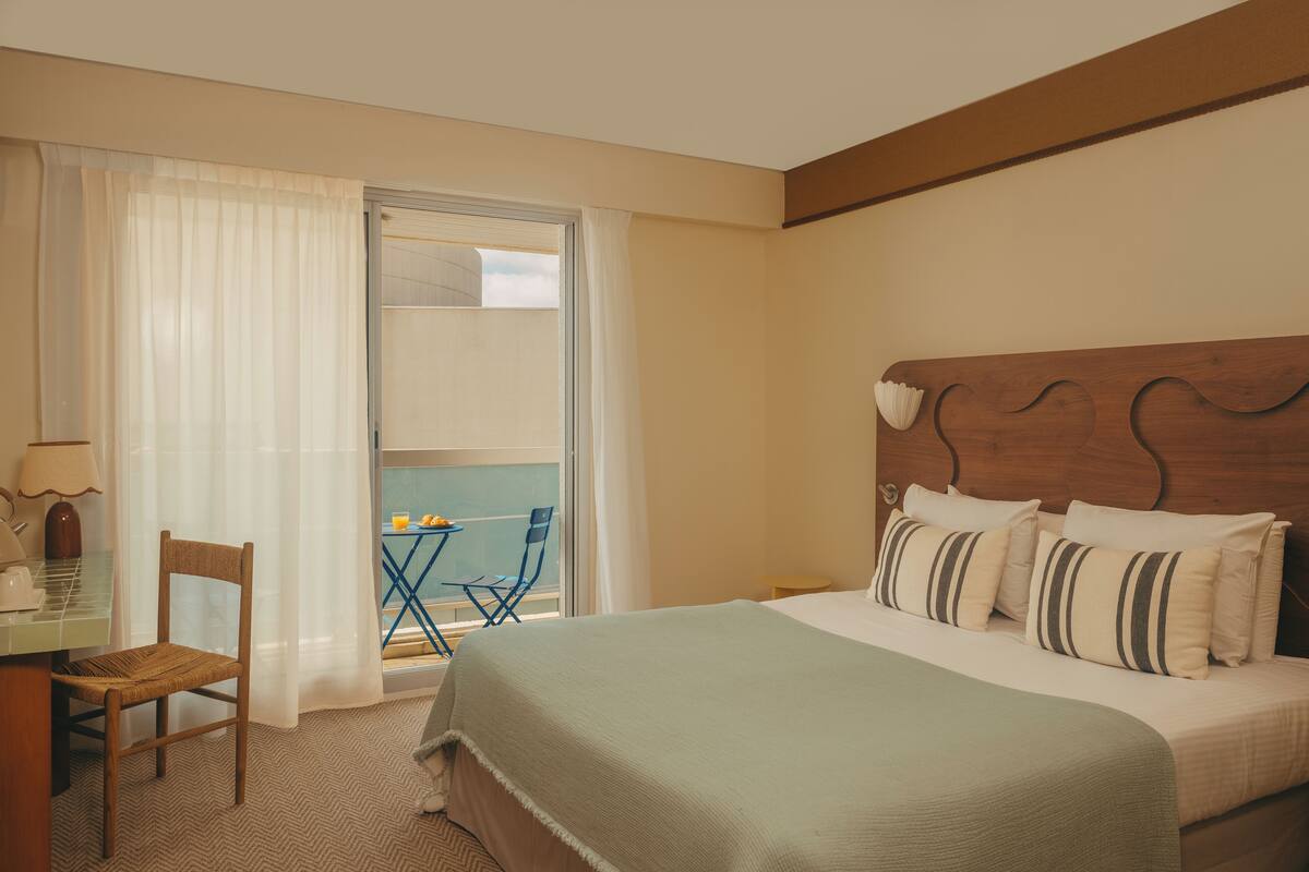 A hotel room with a bed, a desk with a chair, a small table with a chair, and a view through a window.