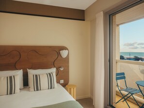 A hotel room with a wooden headboard, a bed with white bedding, a small round table, and a balcony with blue chairs. - Hôtel Coeur Marin (Les Sables-d'Olonne)