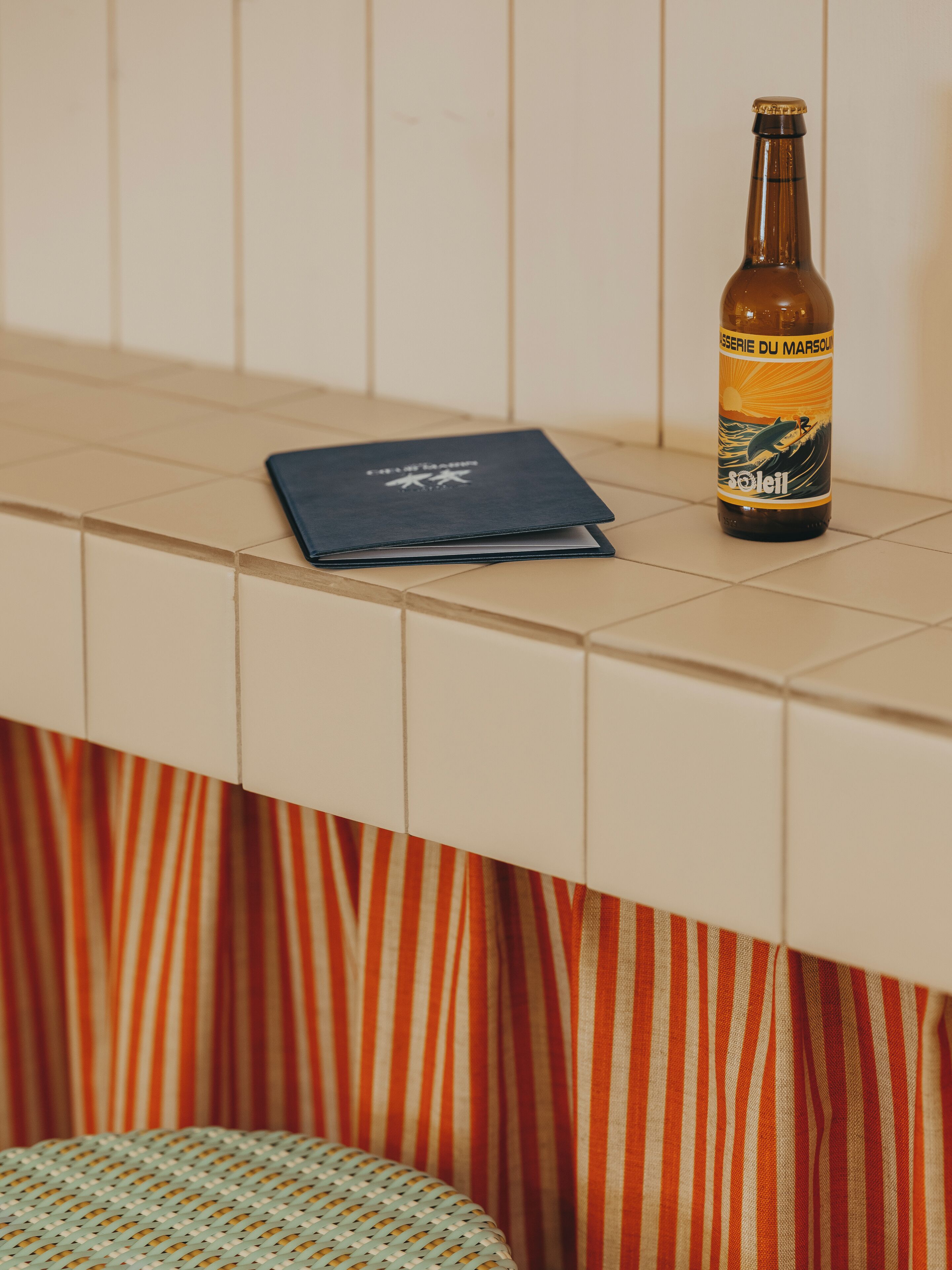 A bottle of beer, a notebook, and a striped curtain on a tiled surface.
