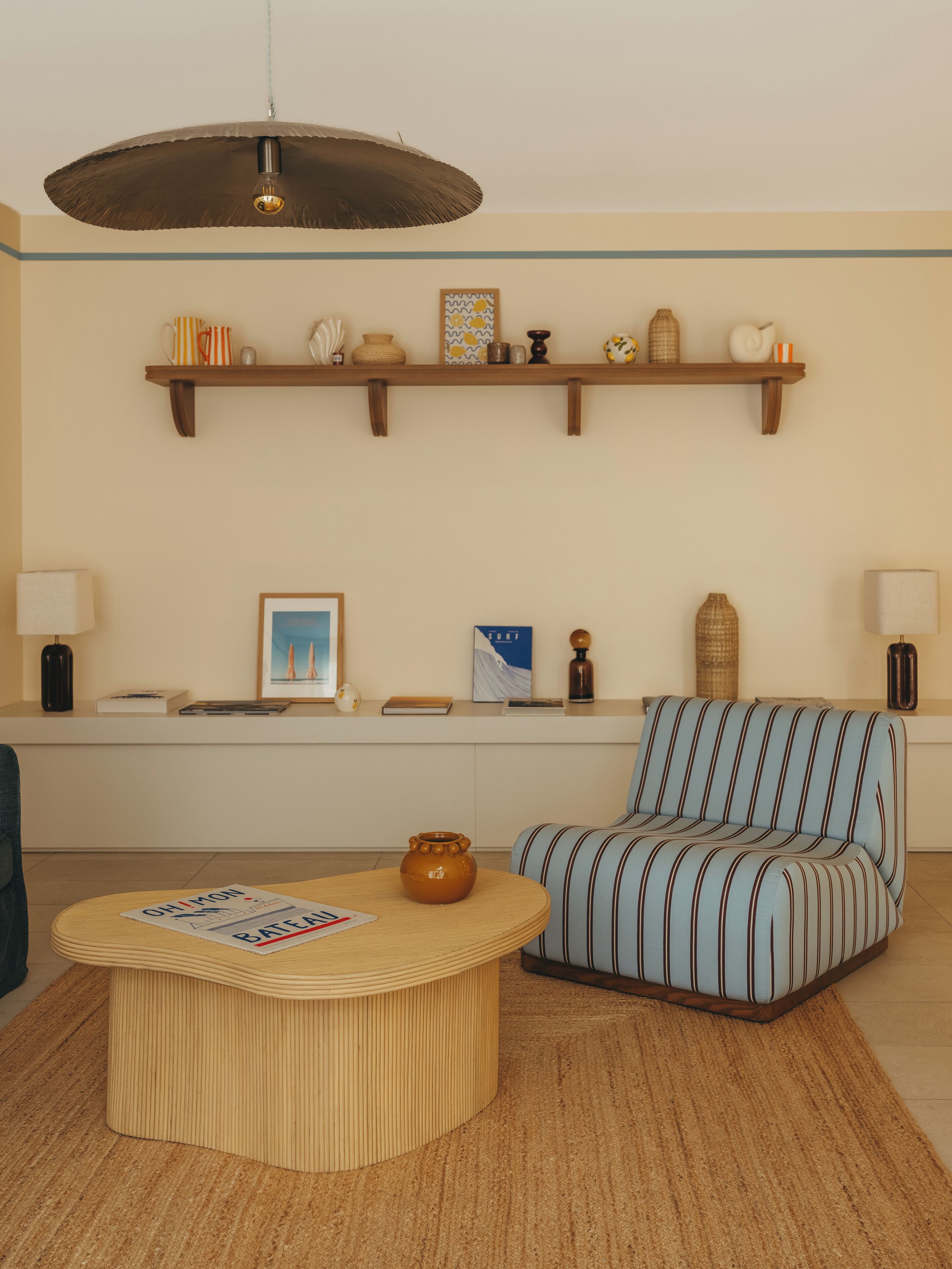 Un salon avec un canapé à rayures, une table basse ronde, un luminaire suspendu et une étagère ornée d’objets décoratifs.