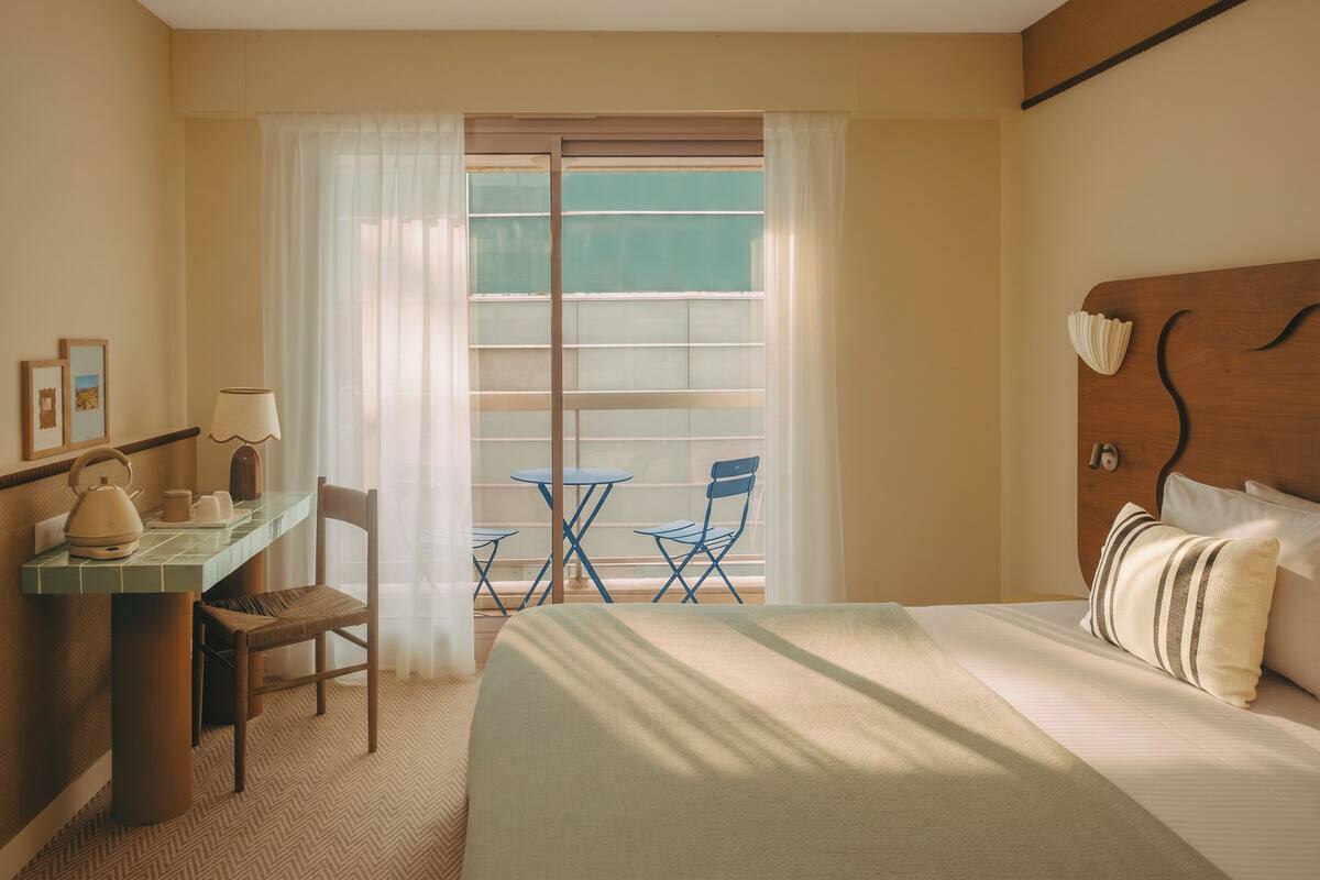 A hotel room with a bed, a desk with a lamp, a chair, a small table, and a view of the outside through a window with curtains.