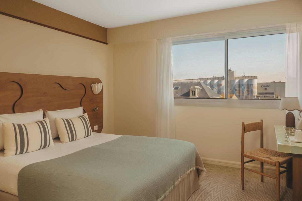 A hotel room with a bed, a chair, a table, and a window with a city view.