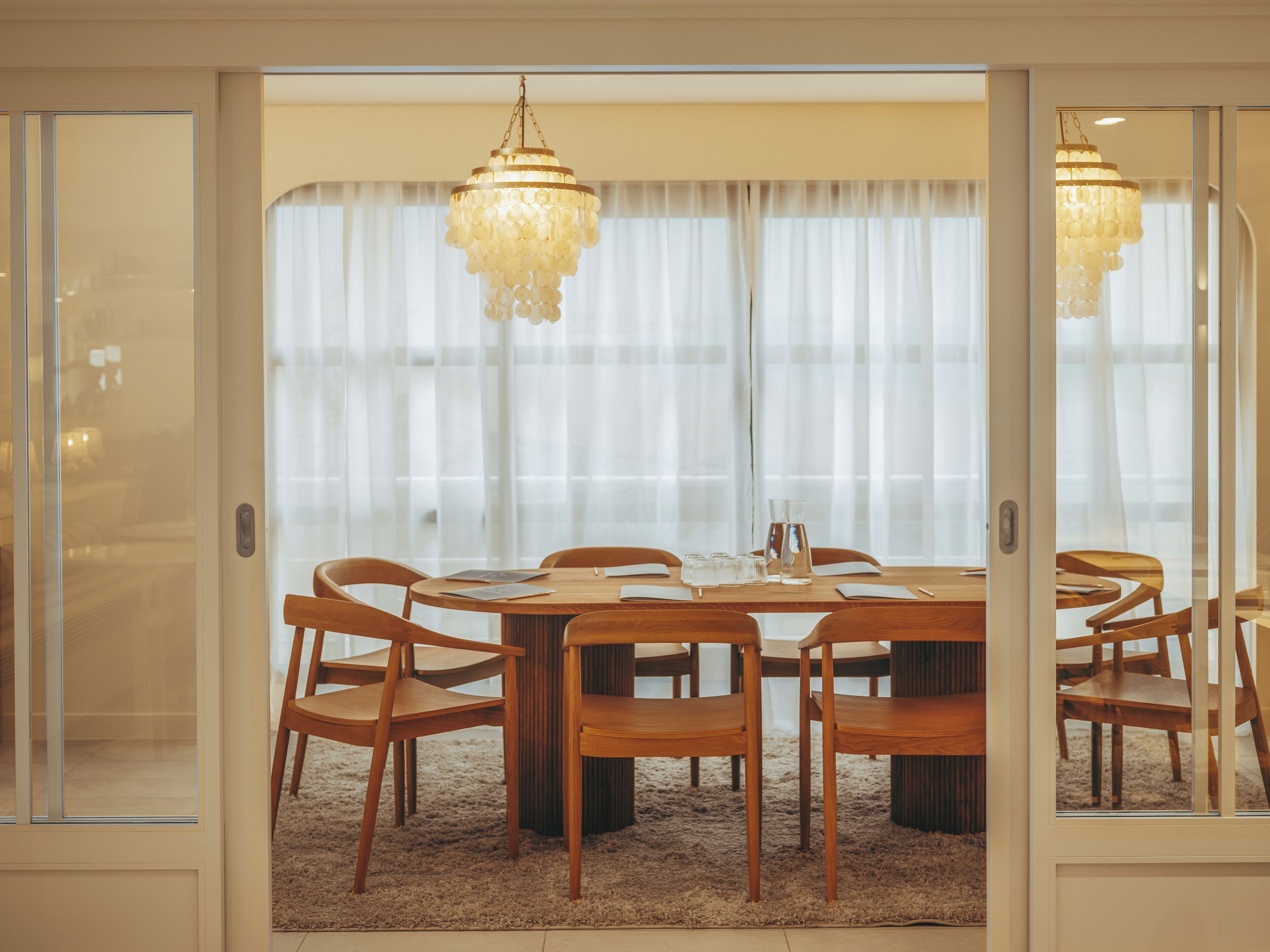 A dining area with a round table, wooden chairs, a chandelier, and a view through a glass door.