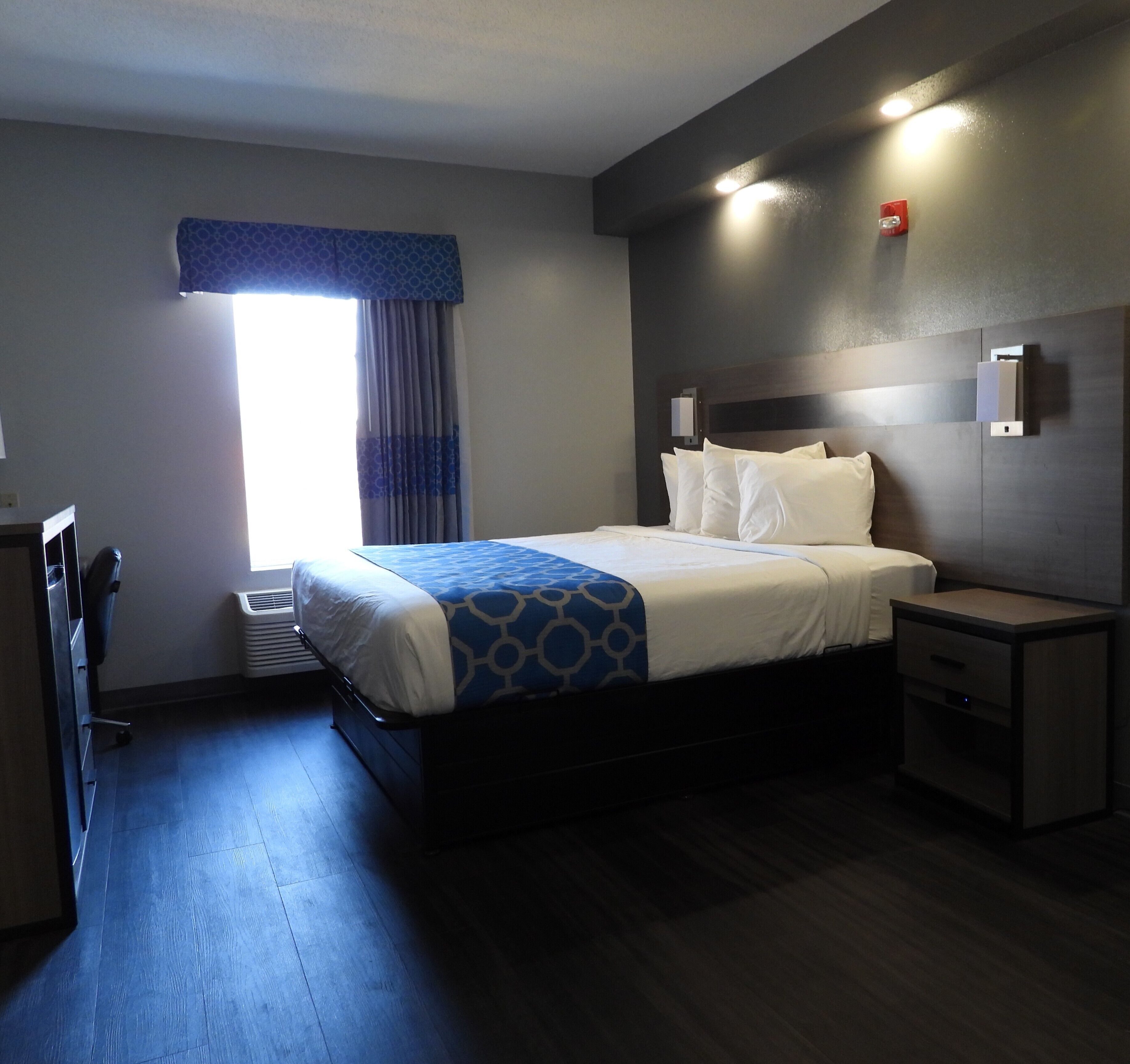 Una habitación de hotel con cama, mesitas de noche, un calentador, una ventana con cortinas, y una lámpara fijada a la pared.