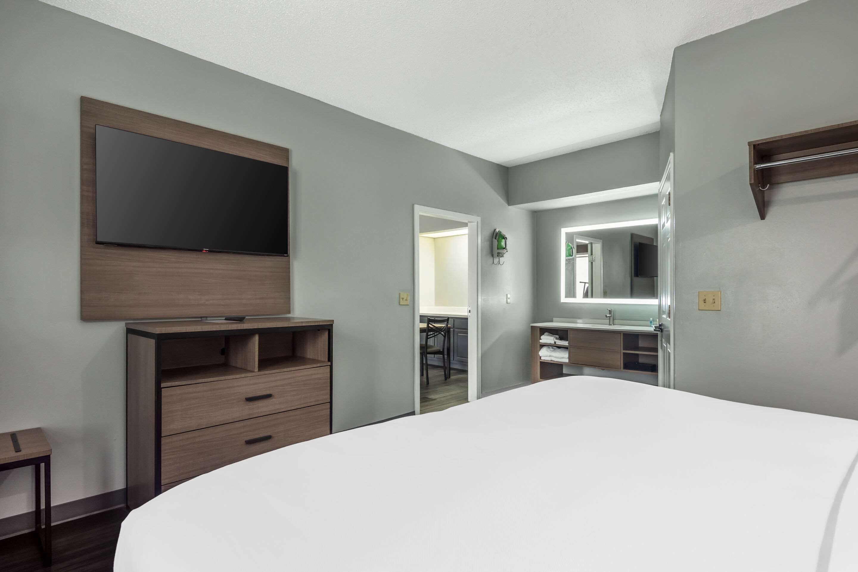 Une chambre d’hôtel moderne avec un grand lit, une commode en bois, un téléviseur à écran plat fixé au mur, une salle de bain avec un miroir et une petite table.