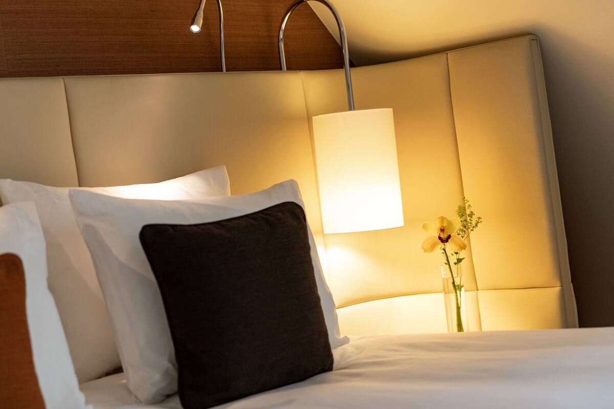 Una cama bien hecha con almohadas blancas y marrones, una lámpara de noche y un florero con flores.