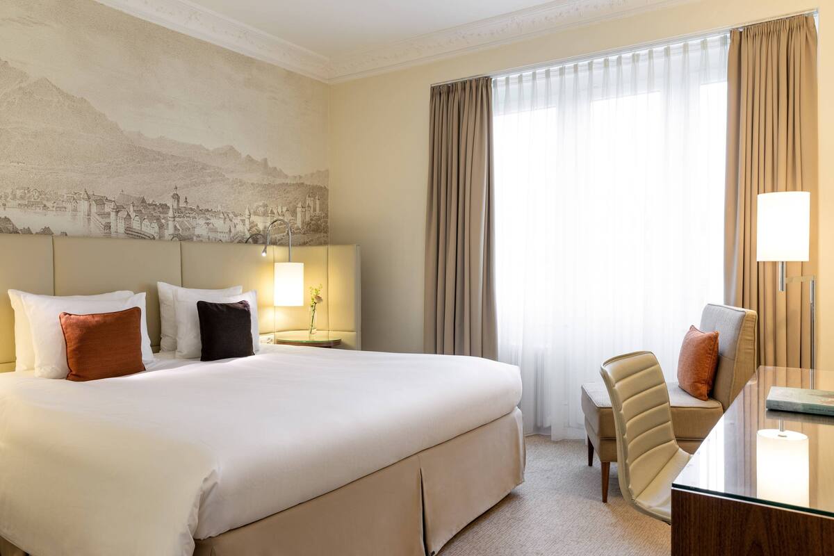 A hotel room with a large bed, a desk, a chair, and a view of a cityscape through the window.