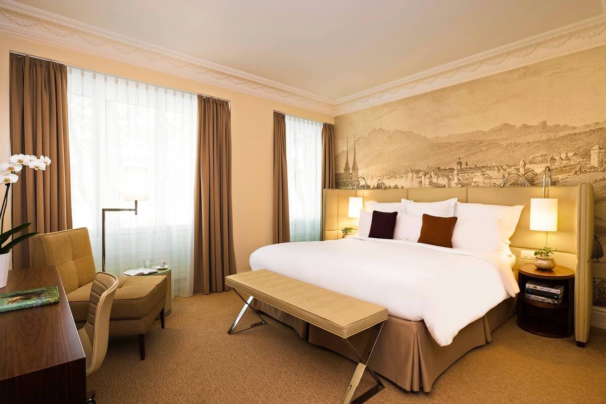 A hotel room with a large bed, a desk, a chair, a bench, and a nightstand with a lamp.