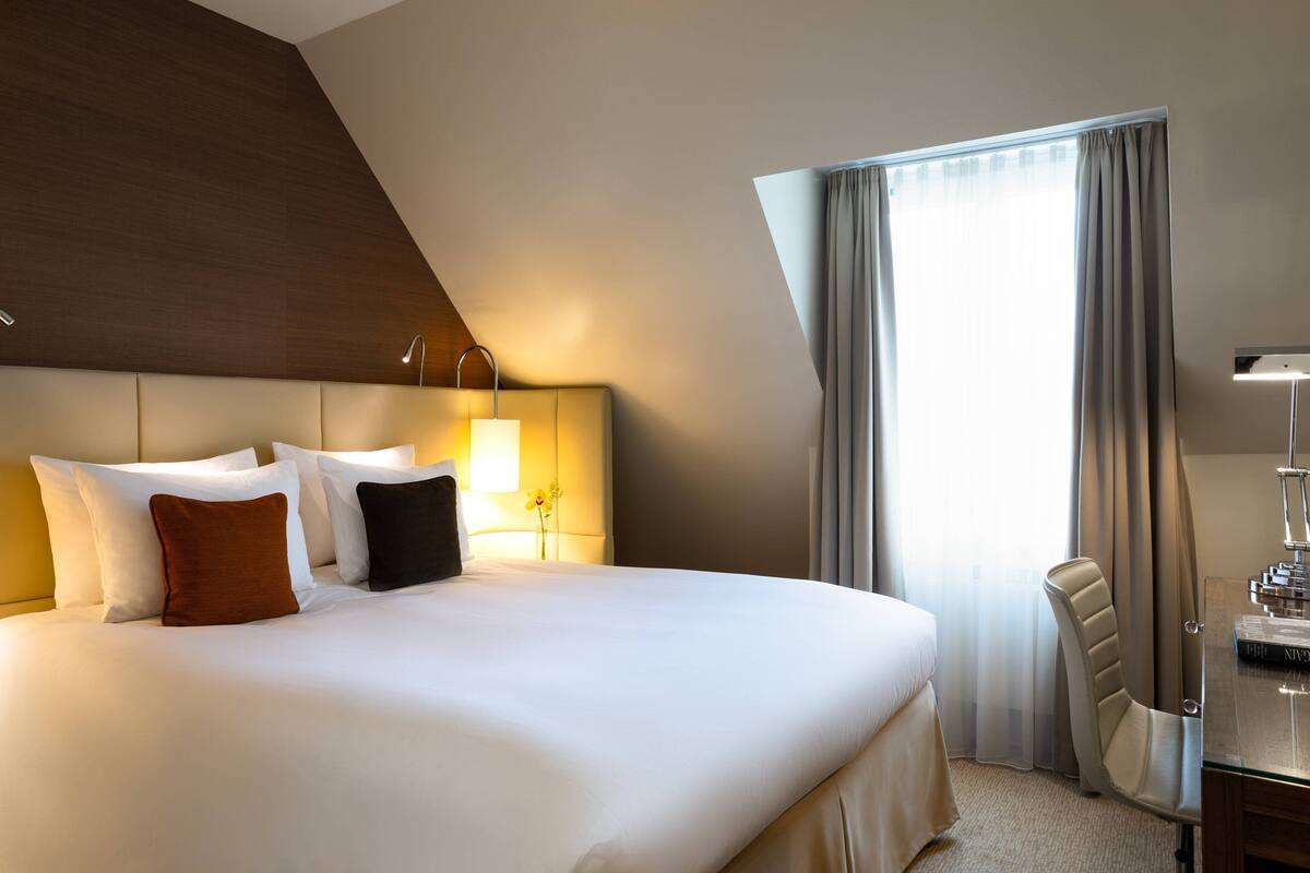 A hotel room with a large bed, a desk with a lamp, and a window with curtains.