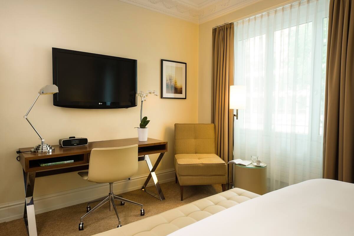 Habitación de hotel con cama, escritorio con computadora, silla, televisor, pequeña mesa con lámpara y ventana con cortinas.