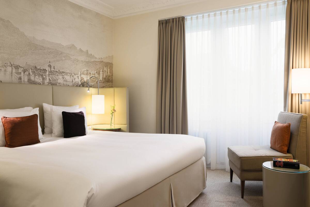 A hotel room with a large bed, a chair, a nightstand with a lamp, and a wall mural.