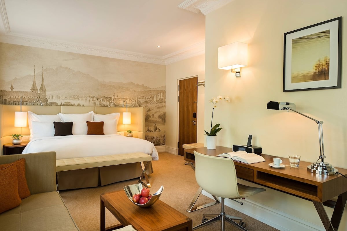 A hotel room with a large bed, a desk with a chair, a small table with a bowl of fruit, and a wall mural.