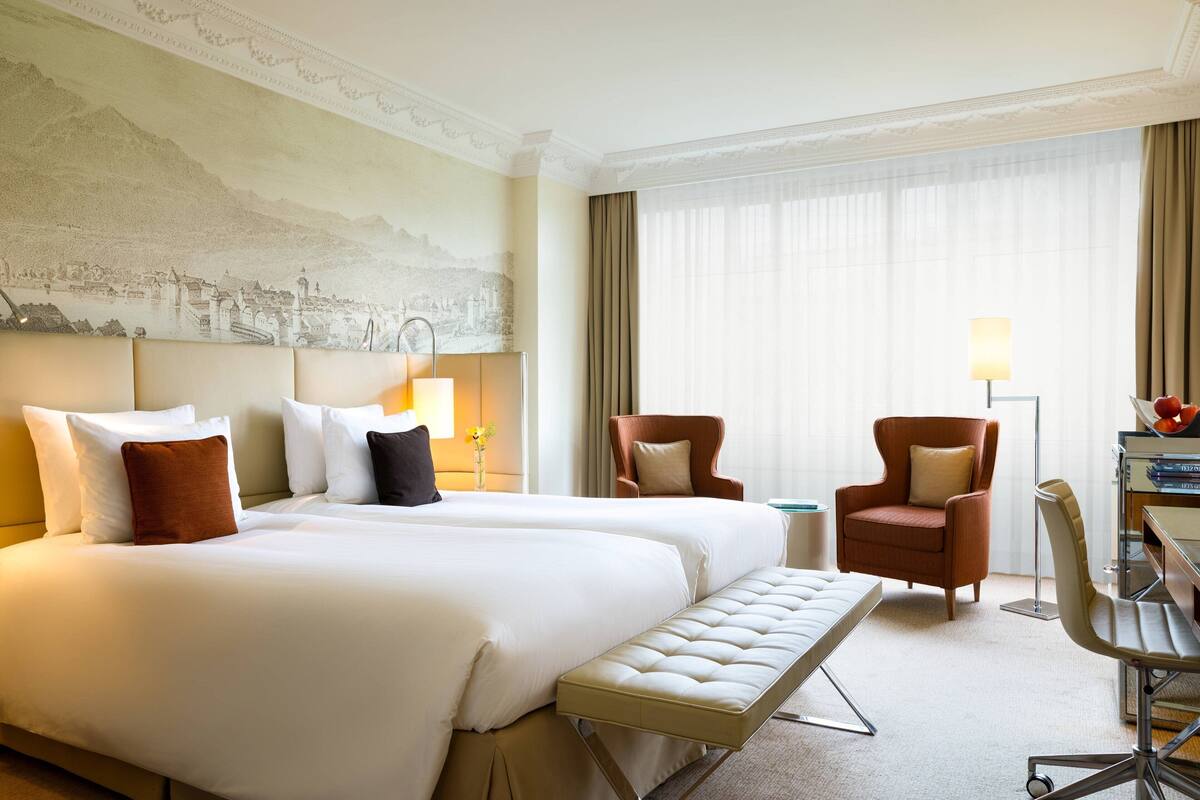 A hotel room with two beds, a bench, a desk, and a chair.