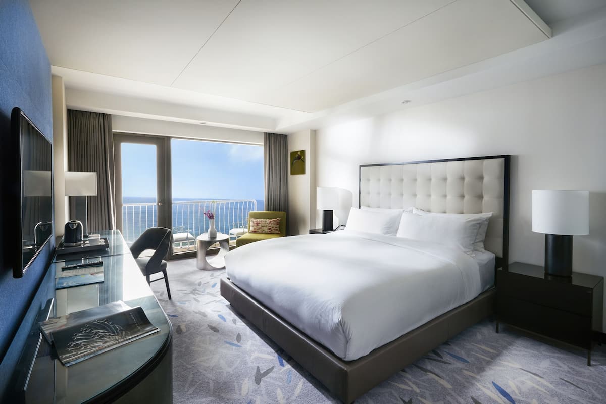 Quarto de hotel moderno com uma cama grande, uma escrivaninha com computador, uma cadeira e vista para o oceano através de amplas janelas.