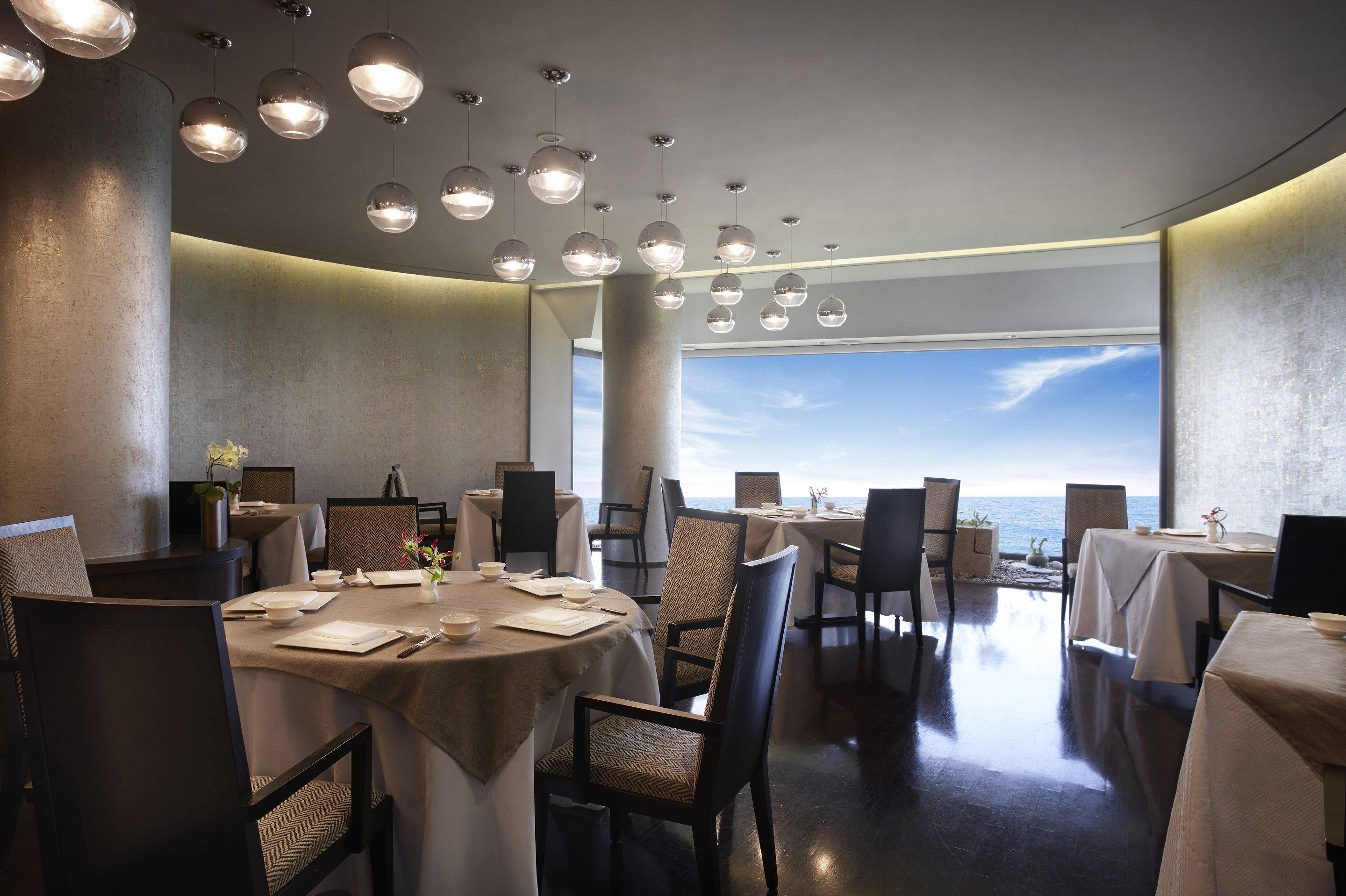 Un restaurante con mesas redondas, sillas, luces esféricas colgantes y vistas al mar.