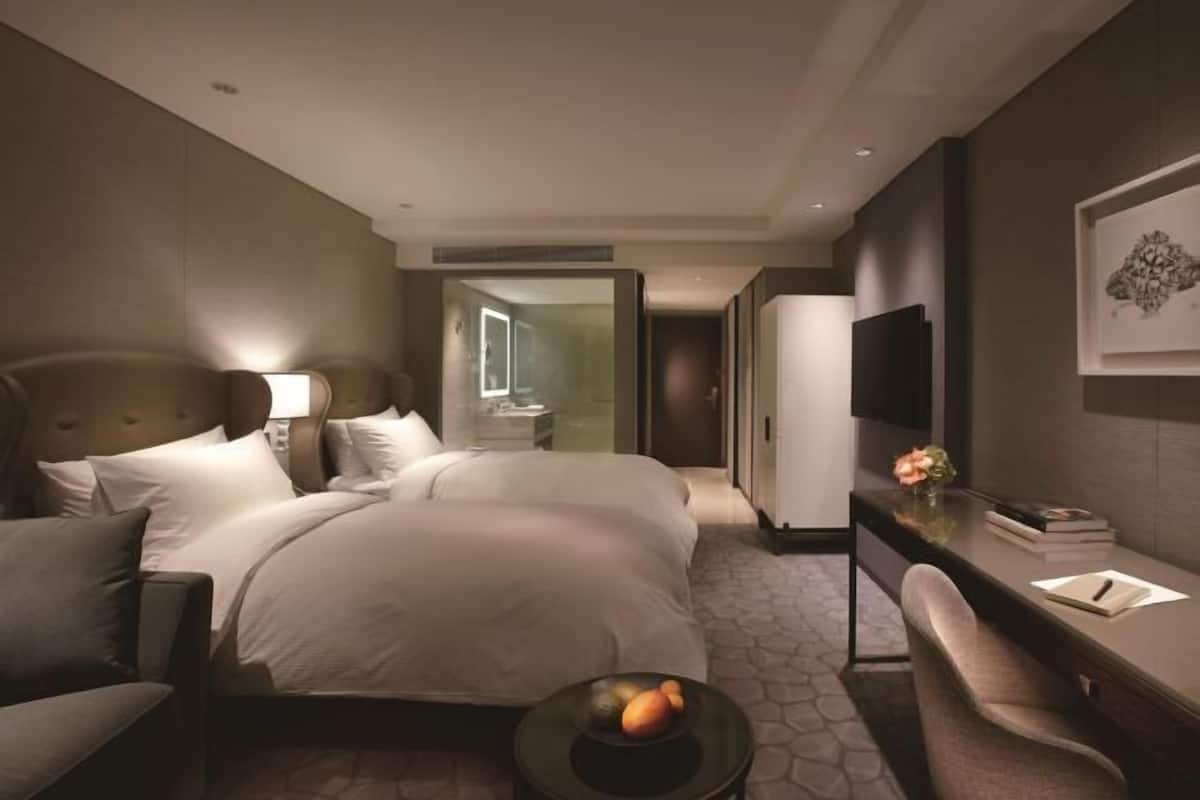 Quarto de hotel moderno com uma cama grande, uma mesa de escritório com cadeira, televisão e uma pequena mesa com frutas.