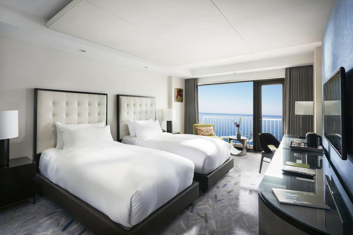 Quarto de hotel com duas camas, uma escrivaninha, uma cadeira e uma grande janela com vista para o mar.