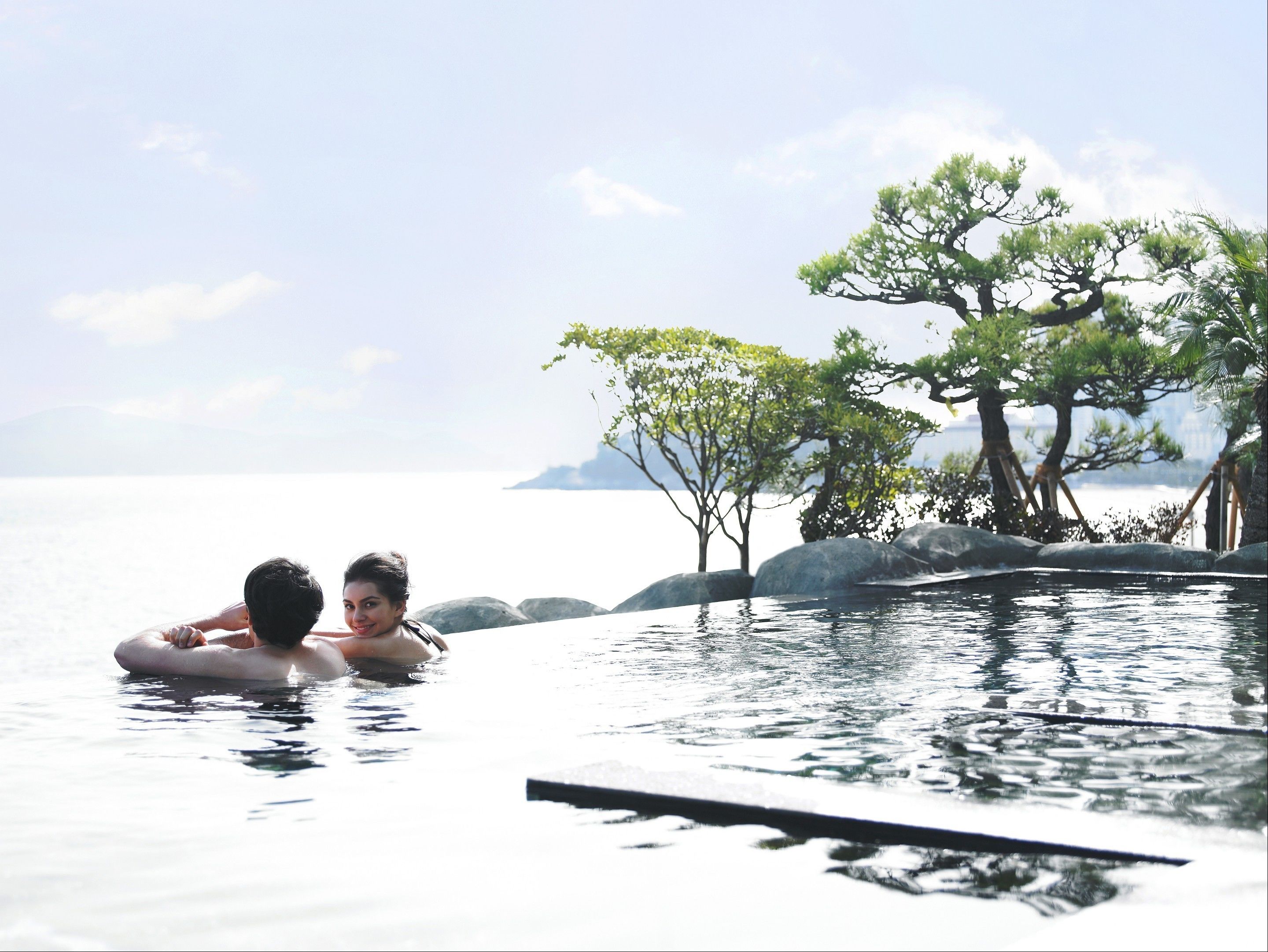 A couple in a clear, calm pool with a view of the sea and trees.