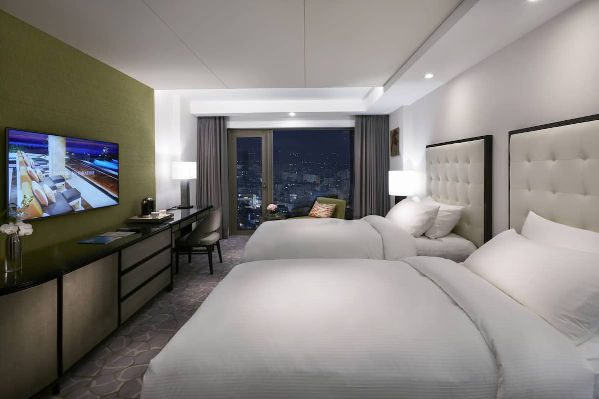 Quarto de hotel com duas camas, uma mesa de escritório, televisão e vista para a cidade à noite.