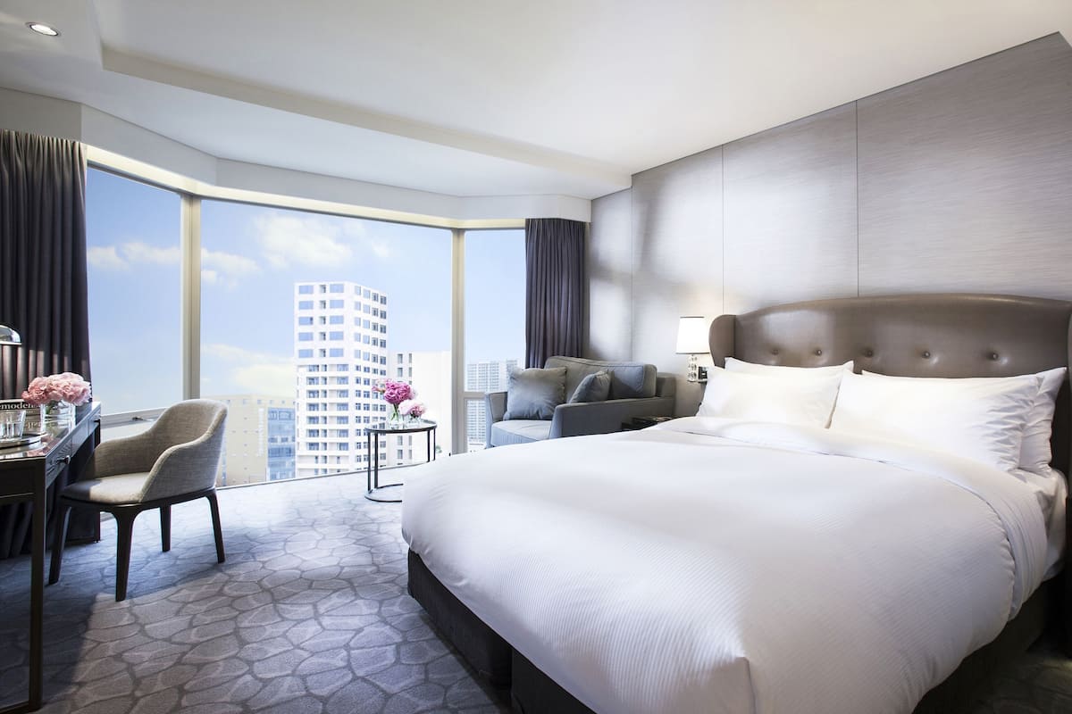 Quarto de hotel moderno com uma cama grande, uma mesa com cadeira, uma mesinha com flores e vista para a cidade.