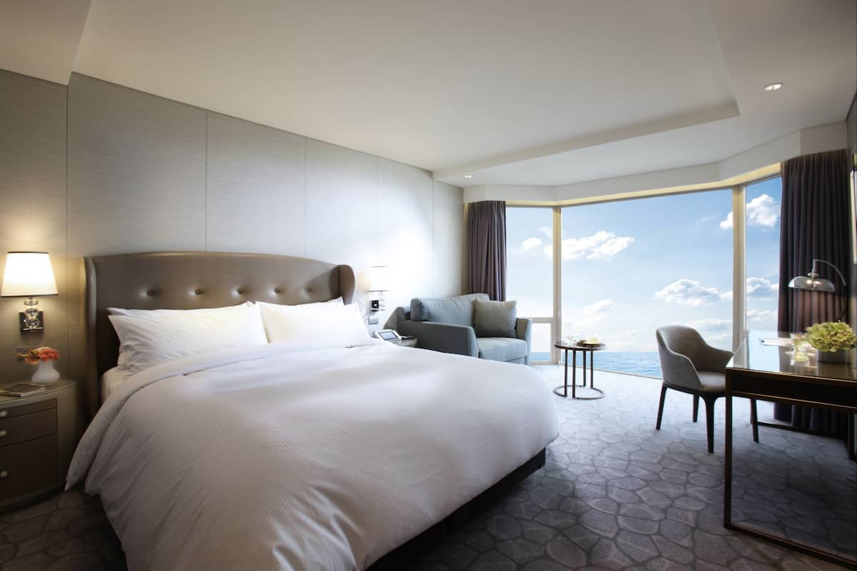 Quarto de hotel moderno com cama grande, escrivaninha, cadeira e vista para o mar.