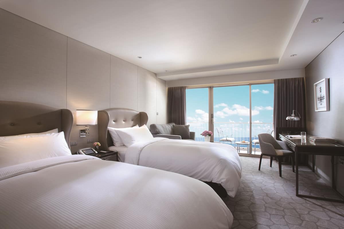 Quarto de hotel com duas camas, uma escrivaninha, uma cadeira e uma janela grande com vista para a cidade.