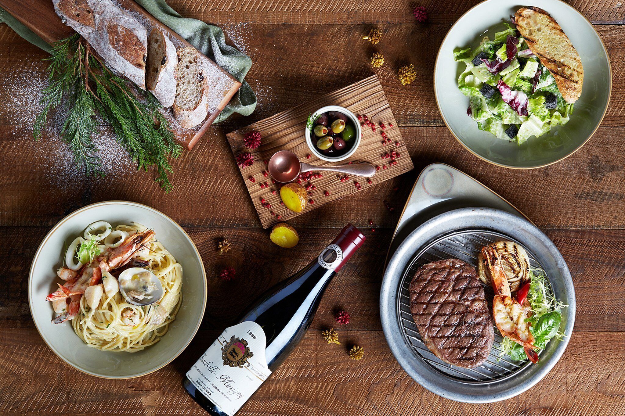 Una mesa de madera con una variedad de platos, como pasta con mariscos, un bistec a la parrilla y una ensalada, además de pan y una botella de vino.