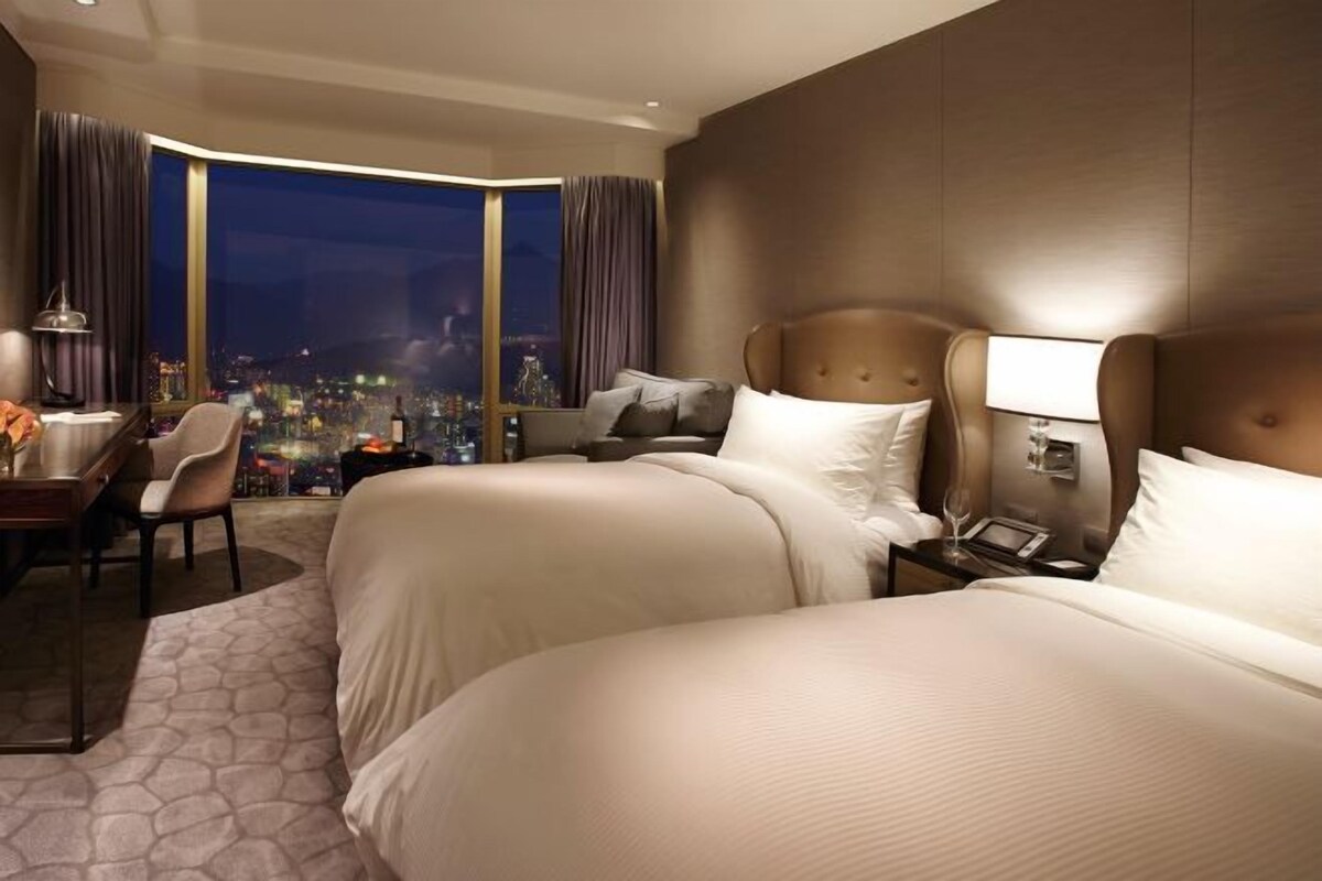 A hotel room with two beds, a desk, a chair, a nightstand, and a view of a cityscape at night.