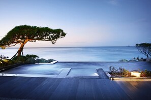 Uma piscina infinita tranquila com vista para o oceano, rodeada por rochas e uma árvore bonsai.