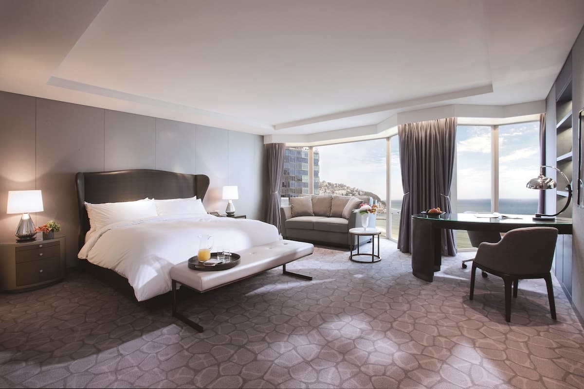 Quarto de hotel com cama, sofá, escrivaninha e cadeira. Possui uma janela grande com cortinas e vista para edifícios.