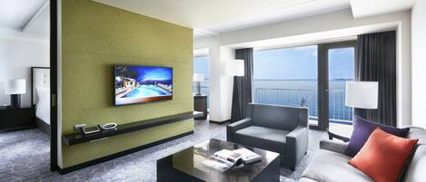 A modern hotel room with a large window, a flat-screen TV, a sofa, a coffee table, and a small side table.