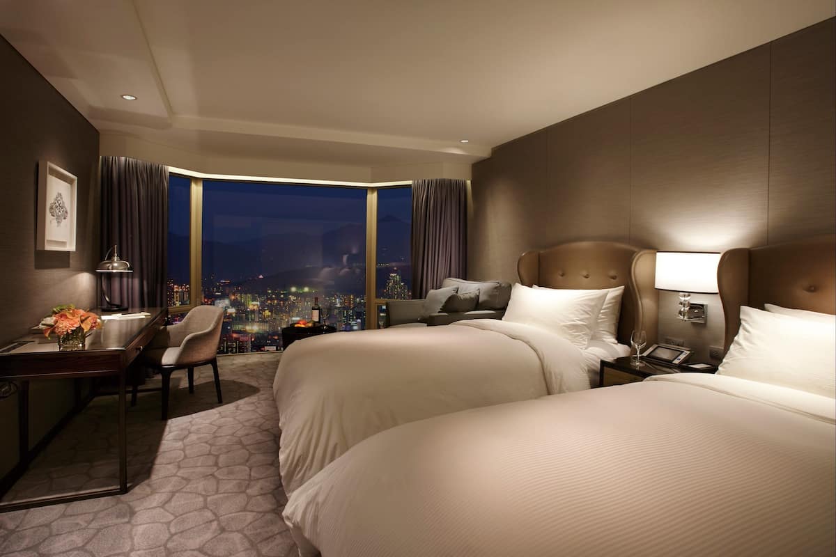 Quarto de hotel com duas camas, uma escrivaninha, uma cadeira, uma mesa de cabeceira e vista para a cidade à noite.