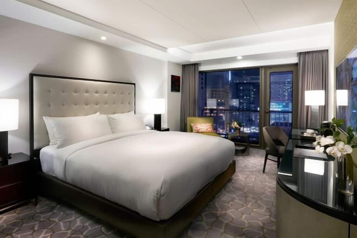 Quarto de hotel com uma cama grande, uma mesa de trabalho e vista da cidade à noite.