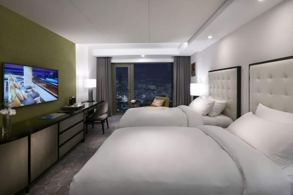 Quarto de hotel com duas camas, uma escrivaninha, uma cadeira, uma televisão e vista para a cidade.