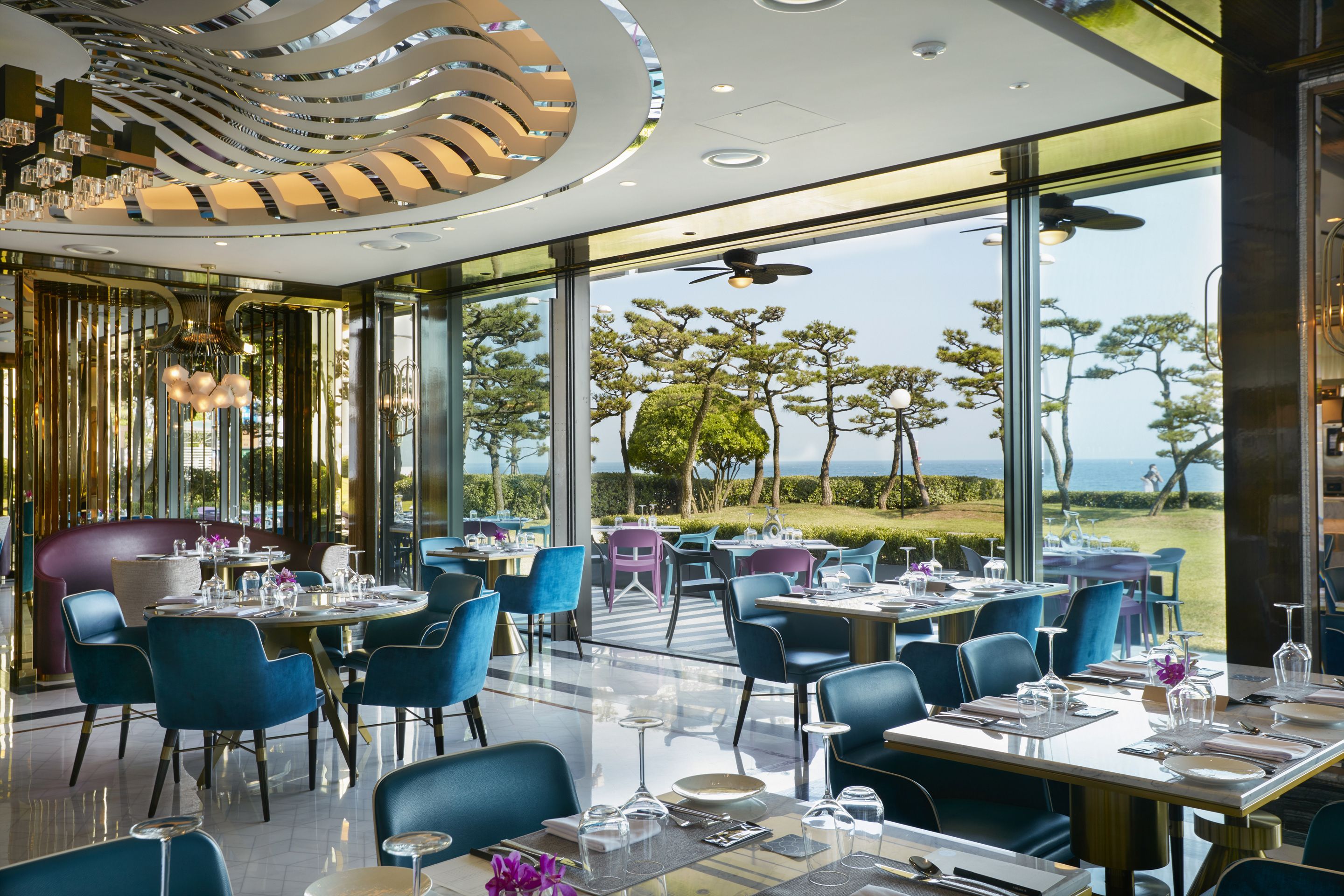 A modern restaurant with a view of the sea, featuring blue chairs, round tables, and a unique ceiling design.