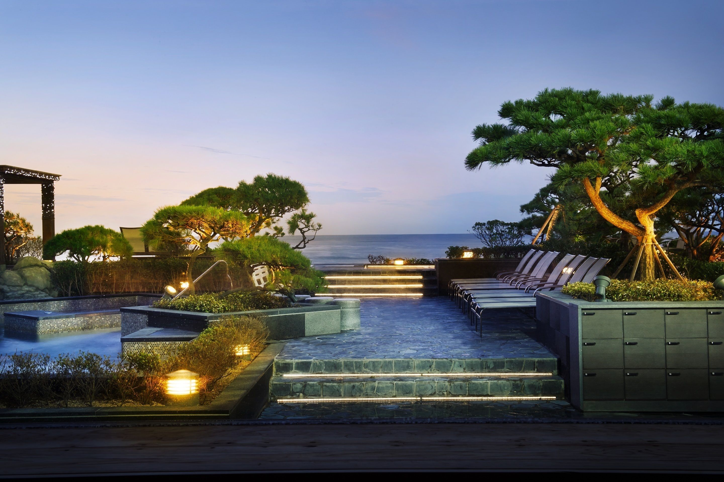 A serene outdoor seating area with steps leading to a beach, featuring a pergola, lounge chairs, and a view of the ocean.