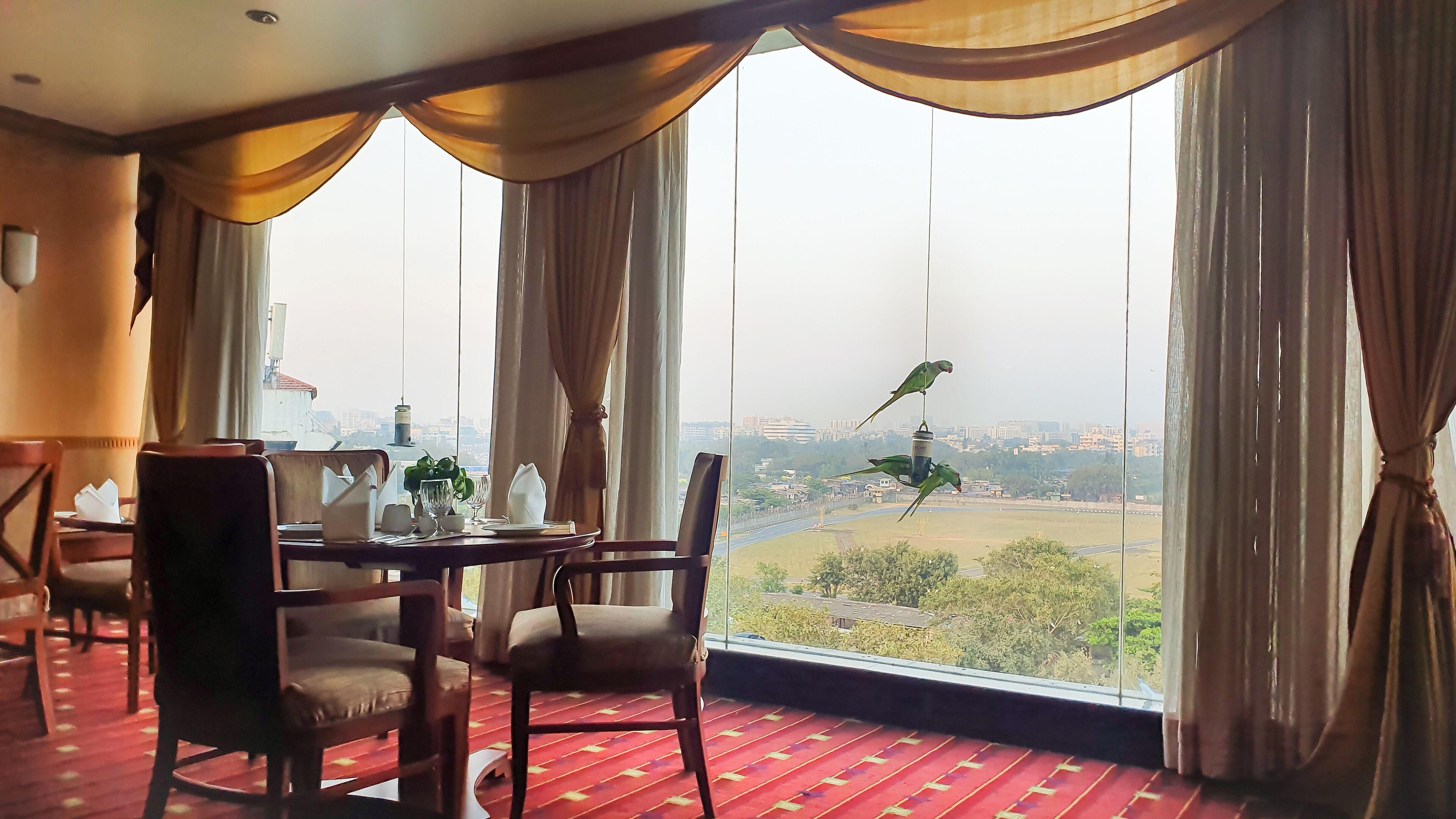 A dining area with a table set for two, large windows with curtains, and a view of a cityscape.