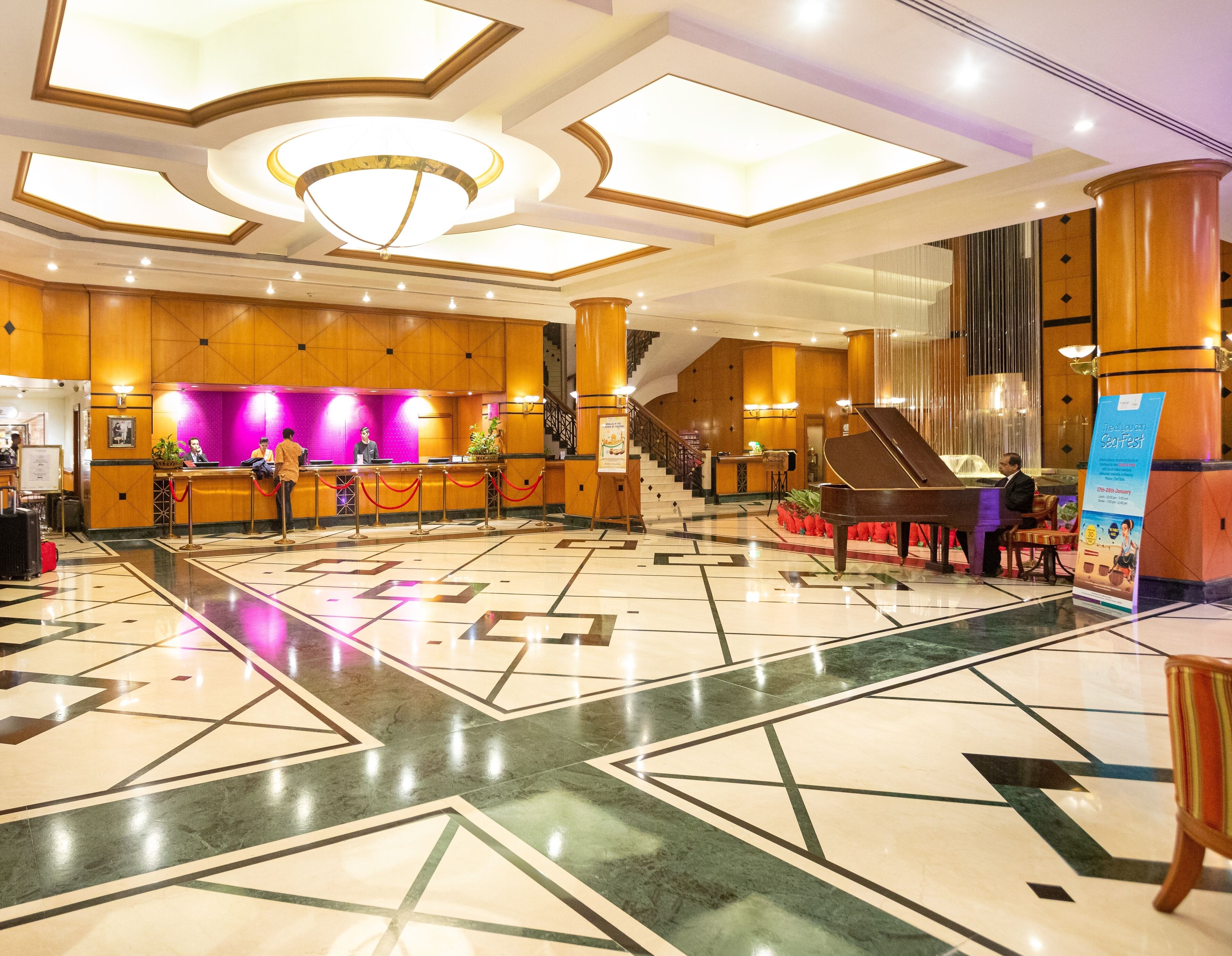 A hotel lobby with a reception desk, a grand piano, a staircase, and a patterned floor.