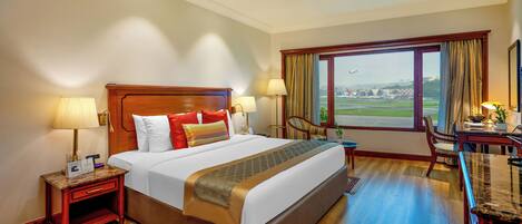 Habitación de hotel con una cama grande, dos mesitas de noche con lámparas, un escritorio con silla y una ventana con vista al aeropuerto.