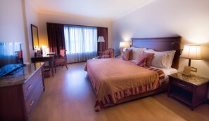 Una habitación de hotel con una cama grande, un escritorio con televisor, una silla y una mesita de noche con lámpara.