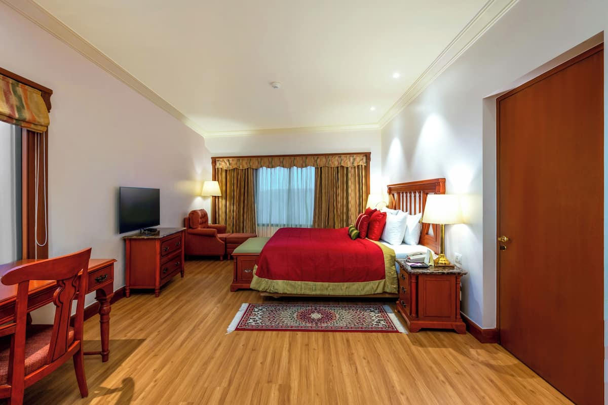 Een hotelkamer met een houten vloer, een bed met rode beddengoed, een bureau met stoel, een televisie en een nachtkastje met lamp.