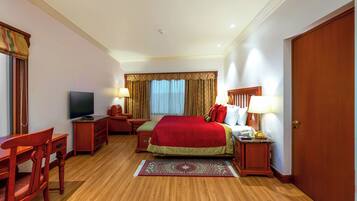 A hotel room with a wooden floor, a bed with red bedding, a desk with a chair, a television, and a nightstand with a lamp.