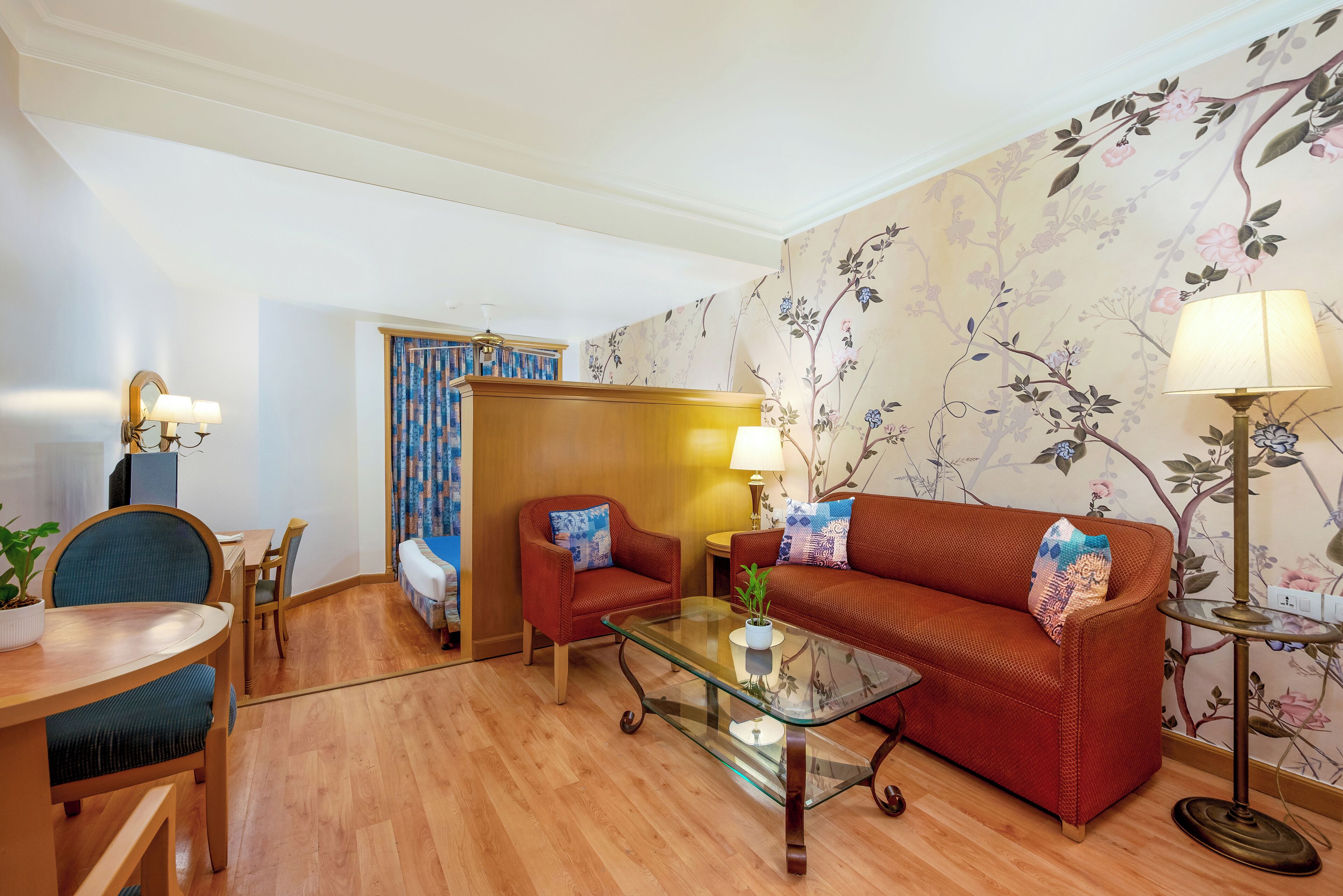 A living room with a wooden floor, a glass coffee table, a red sofa, a blue armchair, a small table with a lamp, and a floral wallpaper.