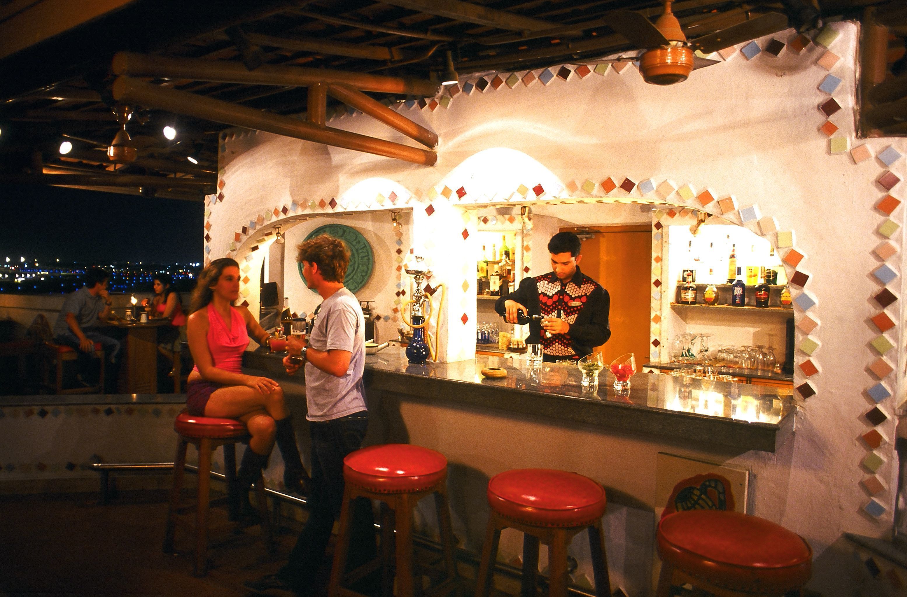 A bar with stools and a counter, a person standing behind the bar, and a city view in the background.